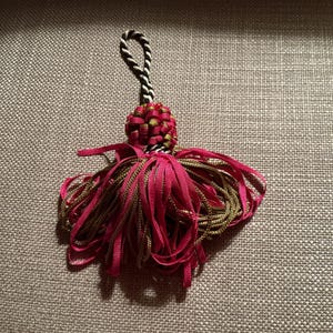 Puede incluir: Una borla decorativa con una parte superior tejida y un lazo para colgar. La borla presenta una combinaci&oacute;n de cintas fucsia y doradas, creando un efecto en capas. La parte superior es una mezcla de rojo y verde, con un cord&oacute;n blanco y negro.