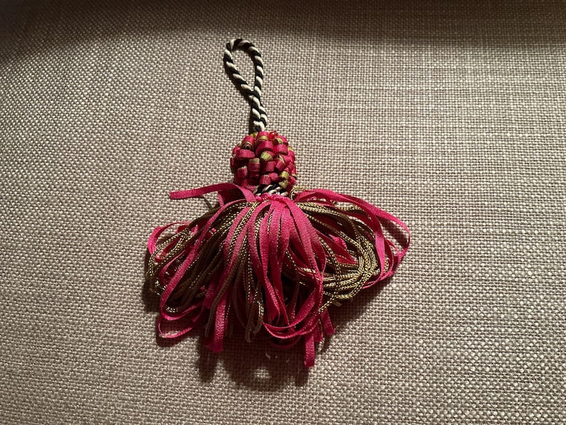 Puede incluir: Una borla decorativa con una parte superior tejida y un lazo para colgar. La borla presenta una combinaci&oacute;n de cintas fucsia y doradas, creando un efecto en capas. La parte superior es una mezcla de rojo y verde, con un cord&oacute;n blanco y negro.