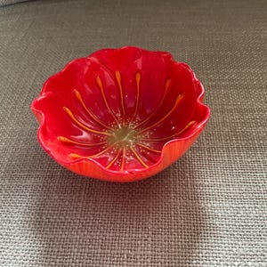 Op de afbeelding: Een felrode, bloemvormige kom met een glanzende afwerking. De kom heeft een gedetailleerd ontwerp met oranje-gele lijnen die vanuit het midden uitstralen, die lijken op bloemblaadjes. De kom is waarschijnlijk gemaakt van glas of een vergelijkbaar materiaal.