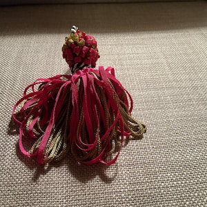 Puede incluir: Un borla decorativa con un grupo de elementos rojos y amarillo-verdosos en la parte superior, que se transforma en una cascada de cintas rojas y doradas. La borla se encuentra sobre un fondo neutro y texturizado.