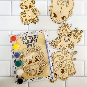May include: Wooden dragon-themed craft kit with paint and a paintbrush. The kit includes several wooden dragon cutouts and a paint palette with red, yellow, blue, green, white, and black paints. The packaging reads "Thank You For Celebrating With Me."