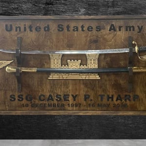 May include: Wooden plaque with the text "United States Army" and "SSG CASEY P. THARP". It features a sword, a saber, and two gold-colored emblems. The plaque is framed in a dark wood.