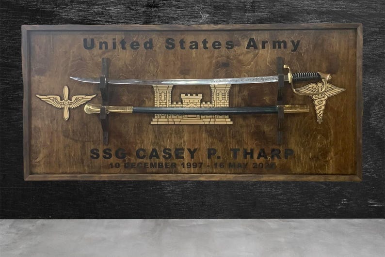May include: Wooden plaque with the text "United States Army" and "SSG CASEY P. THARP". It features a sword, a saber, and two gold-colored emblems. The plaque is framed in a dark wood.