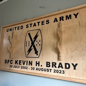 May include: Wooden plaque with the words "UNITED STATES ARMY" and "SFC KEVIN H. BRADY" in black lettering. The plaque features a military emblem and several wooden hooks. The dates "30 JULY 2002 - 30 AUGUST 2023" are also included.