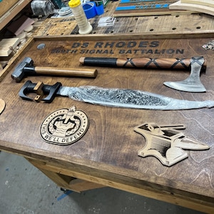 May include: A wooden display featuring a hammer, axe, and a long blade. The display also includes wooden cutouts of a Spartan helmet, a badge with the text "THIS WE'LL DEFEND", and an anvil. The text "DS RHODES 369TH SIGNAL BATTALION" is engraved on the display.