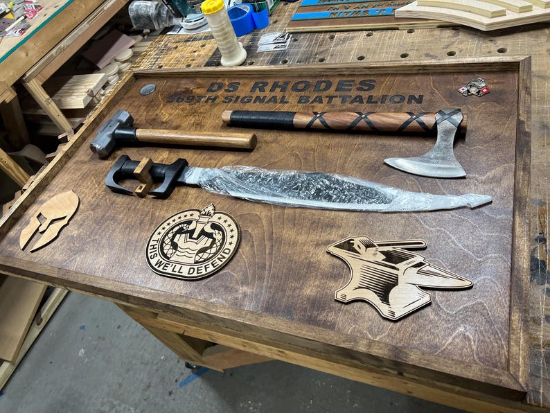 May include: A wooden display featuring a hammer, axe, and a long blade. The display also includes wooden cutouts of a Spartan helmet, a badge with the text "THIS WE'LL DEFEND", and an anvil. The text "DS RHODES 369TH SIGNAL BATTALION" is engraved on the display.