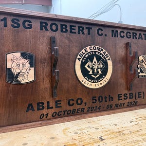 May include: A wooden plaque with the text "1SG ROBERT C. MCGRATH" and "ABLE CO, 50th ESB(E)". It features three emblems and dates. The plaque is stained a dark brown color.