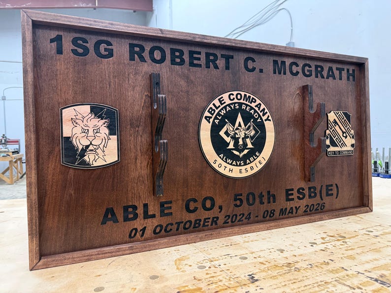 May include: A wooden plaque with the text "1SG ROBERT C. MCGRATH" and "ABLE CO, 50th ESB(E)". It features three emblems and dates. The plaque is stained a dark brown color.