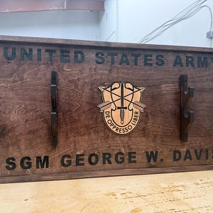 May include: A dark stained wooden plaque with the words "UNITED STATES ARMY" and "SGM GEORGE W. DAVIS" engraved in black. The plaque features a central emblem and two sets of hooks.
