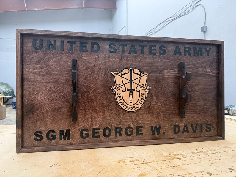 May include: A dark stained wooden plaque with the words "UNITED STATES ARMY" and "SGM GEORGE W. DAVIS" engraved in black. The plaque features a central emblem and two sets of hooks.