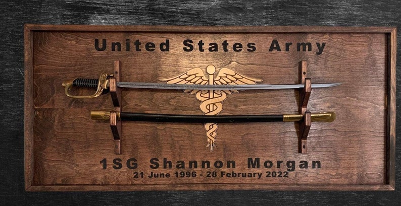 May include: Wooden plaque with a sword and scabbard display. The plaque features the text "United States Army" and "1SG Shannon Morgan 21 June 1996 - 28 February 2022". A medical caduceus is in the center.