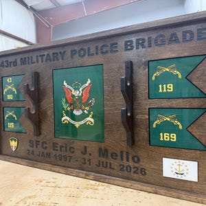 May include: A wooden plaque with the text "43rd MILITARY POLICE BRIGADE". It features green and gold emblems, flags, and the name "SFC Eric J. Mello" with dates. The plaque is brown with a dark border and has a glossy finish.