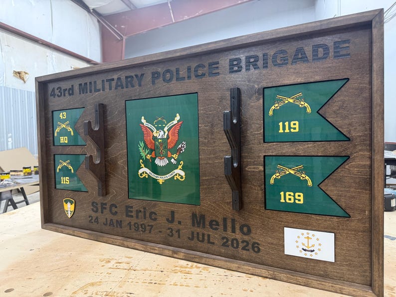 May include: A wooden plaque with the text "43rd MILITARY POLICE BRIGADE". It features green and gold emblems, flags, and the name "SFC Eric J. Mello" with dates. The plaque is brown with a dark border and has a glossy finish.