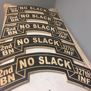 May include: Five wooden signs with black backgrounds and gold lettering. The signs read "2nd BN NO SLACK 327th INF".
