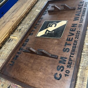 May include: A dark stained wooden plaque with the text "UNITED STATES ARMY" and "CSM STEVEN WALKER 10 SEPTEMBER 2020 - 10 SEPTEMBER 2023". The plaque features a military emblem with the number 75 and cutouts for displaying items.