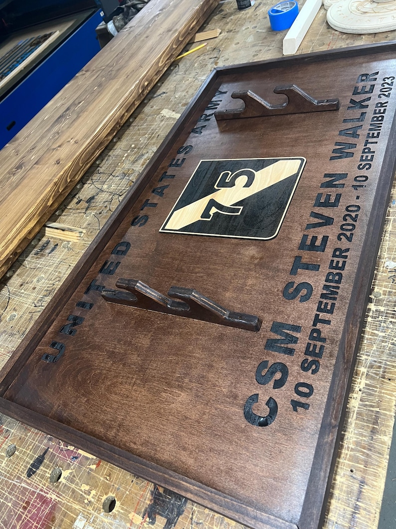 May include: A dark stained wooden plaque with the text "UNITED STATES ARMY" and "CSM STEVEN WALKER 10 SEPTEMBER 2020 - 10 SEPTEMBER 2023". The plaque features a military emblem with the number 75 and cutouts for displaying items.
