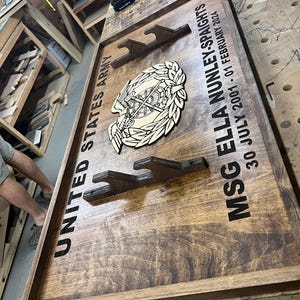 May include: Wooden plaque with the text "UNITED STATES ARMY" and "MSG ELLA NUNLEY-SPAIGHTS 30 JULY 2001 - 01 FEBRUARY 2024". Features a wooden emblem and three gun mounts. The plaque is stained a dark brown color.
