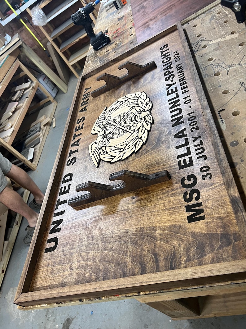 May include: Wooden plaque with the text "UNITED STATES ARMY" and "MSG ELLA NUNLEY-SPAIGHTS 30 JULY 2001 - 01 FEBRUARY 2024". Features a wooden emblem and three gun mounts. The plaque is stained a dark brown color.