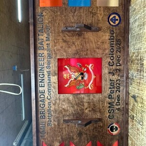 May include: Wooden plaque with a dark frame, featuring various military insignia and text. Includes red, orange, blue, and gold colored markers with numbers and letters. The plaque commemorates the 104th Brigade Engineer Battalion.