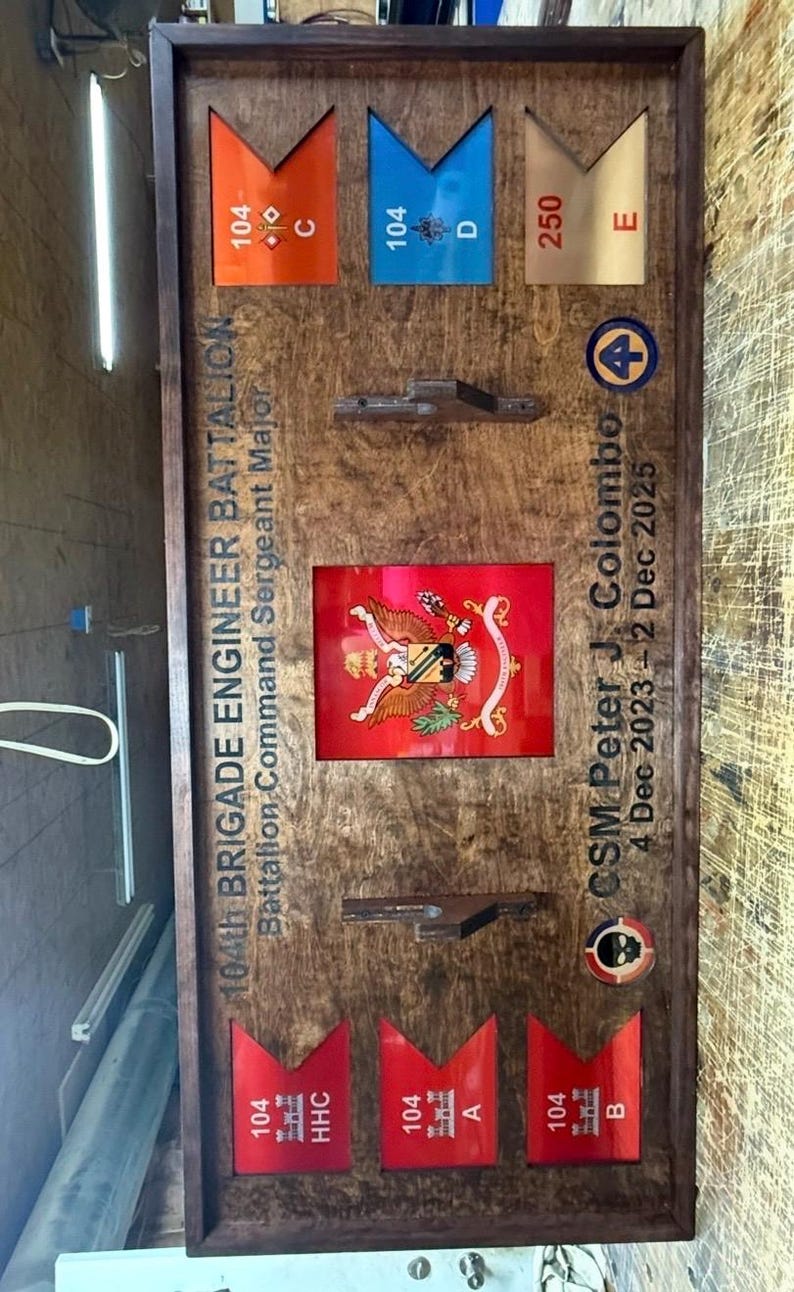 May include: Wooden plaque with a dark frame, featuring various military insignia and text. Includes red, orange, blue, and gold colored markers with numbers and letters. The plaque commemorates the 104th Brigade Engineer Battalion.