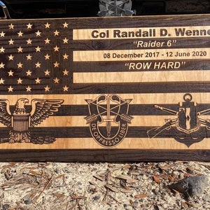 May include: A wooden American flag with a brown and light wood finish. The flag is decorated with a military insignia, the text "Col Randall D. Wenner", "Raider 6", "08 December 2017 - 12 June 2020", and "ROW HARD".