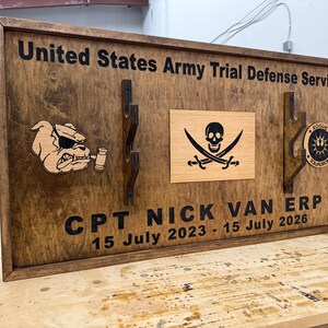 May include: Wooden plaque with the text "United States Army Trial Defense Service" and "CPT NICK VAN ERP 15 July 2023 - 15 July 2026". Features a bulldog, a pirate flag, and a circular emblem. The plaque is framed in dark brown.