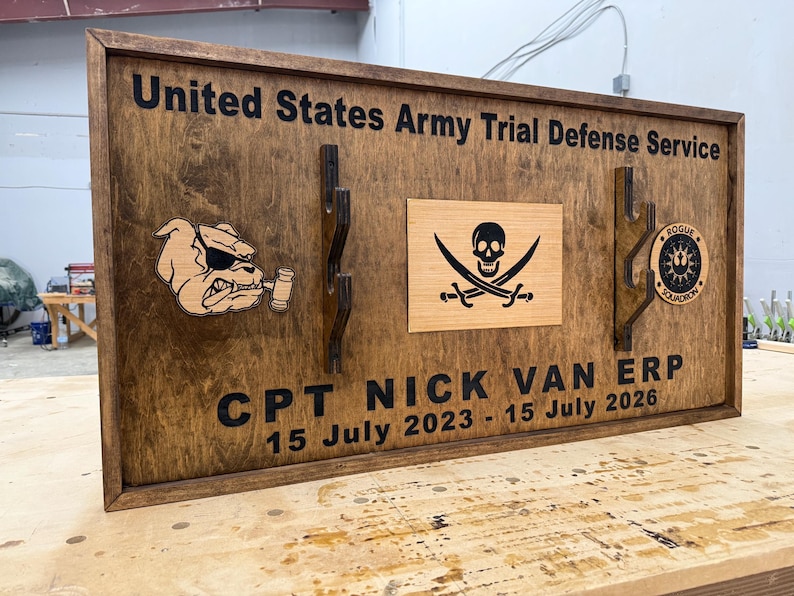 May include: Wooden plaque with the text "United States Army Trial Defense Service" and "CPT NICK VAN ERP 15 July 2023 - 15 July 2026". Features a bulldog, a pirate flag, and a circular emblem. The plaque is framed in dark brown.