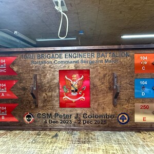 May include: A wooden display case featuring military insignia and text. The case includes various flags with unit designations, a central emblem, and the text "104th BRIGADE ENGINEER BATTALION". It also includes the text "CSM Peter J. Colombo 4 Dec 2023 - 2 Dec 2025".