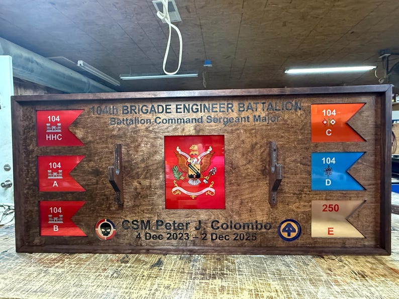 May include: A wooden display case featuring military insignia and text. The case includes various flags with unit designations, a central emblem, and the text "104th BRIGADE ENGINEER BATTALION". It also includes the text "CSM Peter J. Colombo 4 Dec 2023 - 2 Dec 2025".