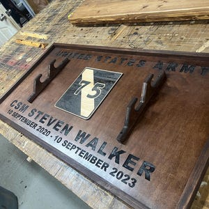 May include: A wooden plaque with the words "UNITED STATES ARMY" and "CSM STEVEN WALKER 10 SEPTEMBER 2020 - 10 SEPTEMBER 2023" engraved in black. It features a patch with the number 75 and two wooden gun holders.