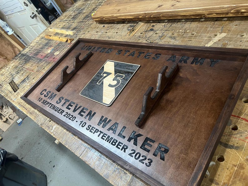 May include: A wooden plaque with the words "UNITED STATES ARMY" and "CSM STEVEN WALKER 10 SEPTEMBER 2020 - 10 SEPTEMBER 2023" engraved in black. It features a patch with the number 75 and two wooden gun holders.