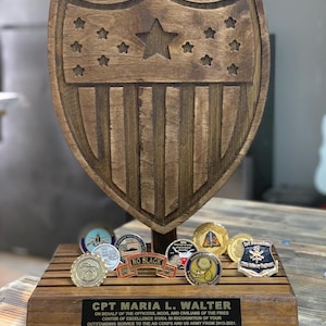 May include: A wooden display featuring a shield design with a star and stripes, holding several military challenge coins. The base includes a plaque with text honoring CPT Maria L. Walter for service in the AG Corps and US Army from 2013-2021.
