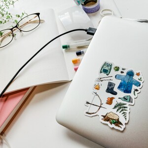 K&ouml;nnte beinhalten: Ein Laptop mit einem Aufkleber mit Aquarellillustrationen von Outdoor-Artikeln, darunter ein Rucksack, ein Mantel, Stiefel und Pfeil und Bogen. Das Bild enth&auml;lt auch Notizb&uuml;cher, Stifte, Brillen und Washi-Tape, alles auf einer wei&szlig;en Oberfl&auml;che.