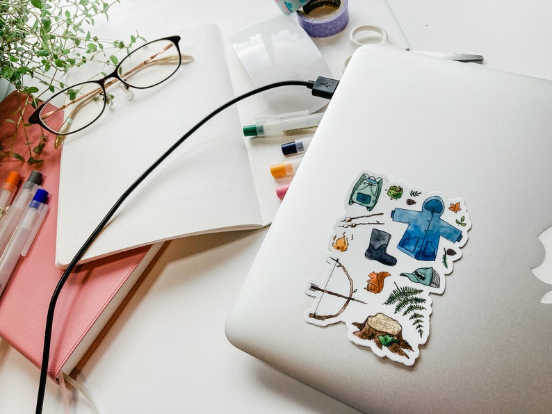 K&ouml;nnte beinhalten: Ein Laptop mit einem Aufkleber mit Aquarellillustrationen von Outdoor-Artikeln, darunter ein Rucksack, ein Mantel, Stiefel und Pfeil und Bogen. Das Bild enth&auml;lt auch Notizb&uuml;cher, Stifte, Brillen und Washi-Tape, alles auf einer wei&szlig;en Oberfl&auml;che.