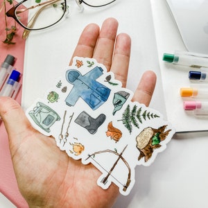 K&ouml;nnte beinhalten: Eine Sammlung von Aquarell-Aufklebern mit Illustrationen zum Thema Outdoor und Camping. Die Aufkleber zeigen eine blaue Jacke, einen Rucksack, Stiefel, Pfeil und Bogen, ein Eichh&ouml;rnchen und ein Lagerfeuer. Die Aufkleber werden in einer Hand gehalten.