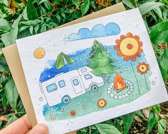 May include: A watercolor illustration on a card depicts a camping scene with a camper, tent, campfire, and flowers. The card is held against a backdrop of green foliage. The artwork features a blue sky, a yellow sun, and a brown envelope.