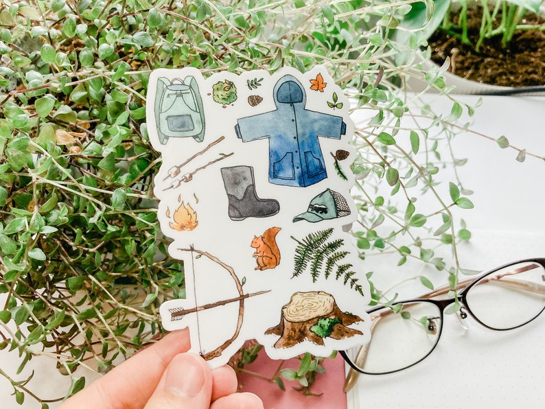 K&ouml;nnte beinhalten: Ein Stickerbogen mit Aquarellillustrationen von Camping- und Outdoor-Abenteuer-Elementen. Enth&auml;lt einen Rucksack, einen blauen Regenmantel, ein Lagerfeuer, einen Pfeil und Bogen, ein Eichh&ouml;rnchen und einen Baumstumpf.