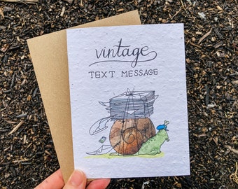 May include: A handmade greeting card with a brown envelope. The card features a whimsical illustration of a snail carrying a stack of papers, with the words "vintage TEXT MESSAGE" in elegant script. The snail is green and brown.