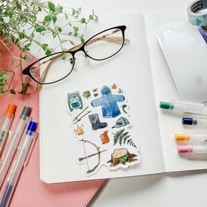 K&ouml;nnte beinhalten: Eine Anordnung von Schreibwaren auf einer wei&szlig;en Oberfl&auml;che, darunter ein Notizbuch, Stifte, eine Brille und Aufkleber mit Camping-Illustrationen. Dekoband und eine Computermaus sind ebenfalls vorhanden, was eine Arbeitsplatzszene schafft.