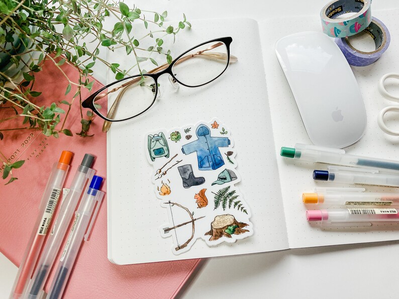 K&ouml;nnte beinhalten: Eine Anordnung von Schreibwaren auf einer wei&szlig;en Oberfl&auml;che, darunter ein Notizbuch, Stifte, eine Brille und Aufkleber mit Camping-Illustrationen. Dekoband und eine Computermaus sind ebenfalls vorhanden, was eine Arbeitsplatzszene schafft.