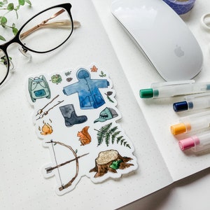 K&ouml;nnte beinhalten: Ein Stickerbogen mit Aquarellillustrationen zum Thema Camping. Enth&auml;lt einen Rucksack, eine Regenjacke, Stiefel, einen Hut, Pfeil und Bogen, Lagerfeuer, Eichh&ouml;rnchen, Farn und einen Baumstumpf. Der Stickerbogen liegt auf einem karierten Notizbuch.