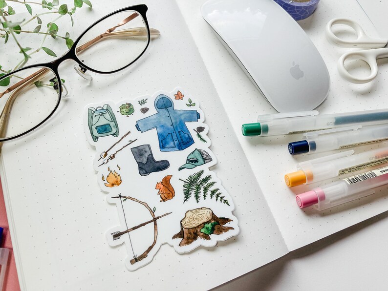 K&ouml;nnte beinhalten: Ein Stickerbogen mit Aquarellillustrationen zum Thema Camping. Enth&auml;lt einen Rucksack, eine Regenjacke, Stiefel, einen Hut, Pfeil und Bogen, Lagerfeuer, Eichh&ouml;rnchen, Farn und einen Baumstumpf. Der Stickerbogen liegt auf einem karierten Notizbuch.