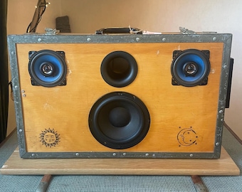 May include: A handmade wooden speaker cabinet with a light brown finish and a metal frame. It has two small blue speakers, a larger black speaker, and a central black speaker. The design includes sun and moon graphics.