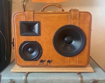 May include: A custom speaker system housed in a vintage brown suitcase. The suitcase has two black speakers, a black horn tweeter, and a handle. Two camel silhouettes are printed on the front. The speaker system is placed on top of a vintage suitcase.