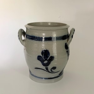 May include: A light gray stoneware crock featuring two side handles and a dark blue hand-painted floral motif on the front. The vessel has a wide circular opening, a thick rim, and horizontal blue bands encircling the upper and lower sections.