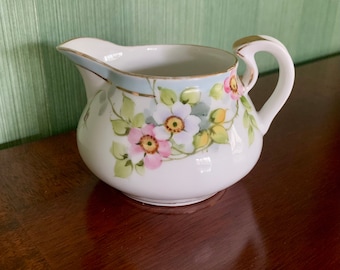 May include: A white porcelain creamer with a floral design. The creamer has a light blue band with gold trim and features pink, white, and yellow flowers with green leaves.