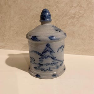 May include: A cylindrical ceramic jar with a matching lid, featuring a light grey base with hand-painted cobalt blue designs. The artwork depicts a stylised evergreen tree, mountains, and birds. A small blue stamp is visible on the lid's edge.