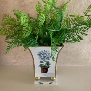 May include: A white square vase with gold-painted edges and four gold spherical feet. The front features a hand-painted illustration of a blue hydrangea in a brown pot. The vase is filled with a lush, vibrant green artificial fern plant.