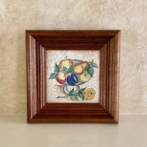 May include: A framed ceramic tile with a hand-painted still life of fruit in a bowl. The fruit includes apples, pears, and plums. The tile has a crackled glaze and is set in a dark wood frame.