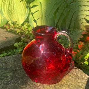May include: A small, round, ruby red glass pitcher with a crackled texture and a clear glass handle. The vessel features a wide, bulbous body that tapers toward a short, open neck, showcasing a vibrant, translucent crimson color throughout.